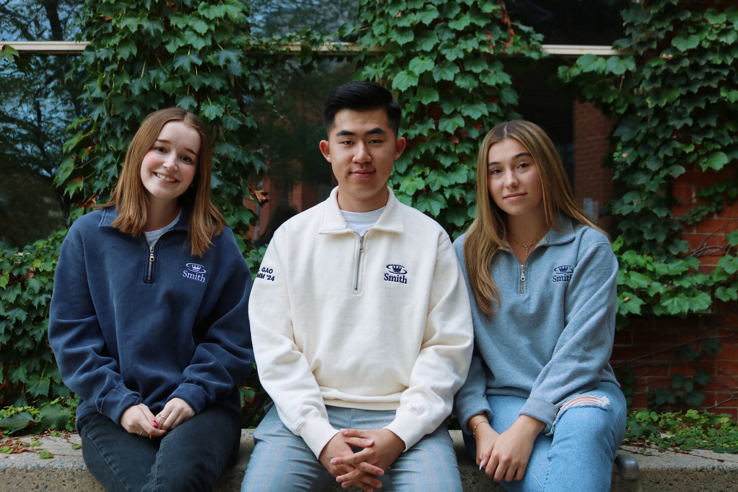 Three young people sitting on a bench in front of a building, wearing Oil Thigh Designs' Smith Quarter Zips.